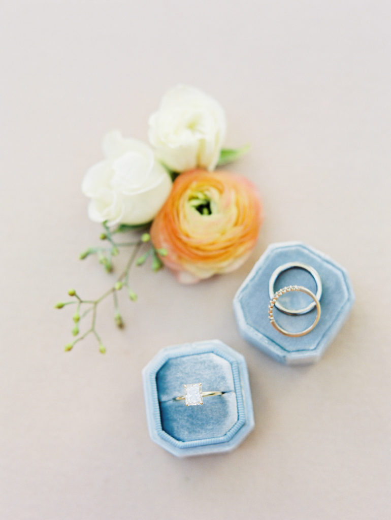 Wedding rings in blue box next to peach and white flowers.