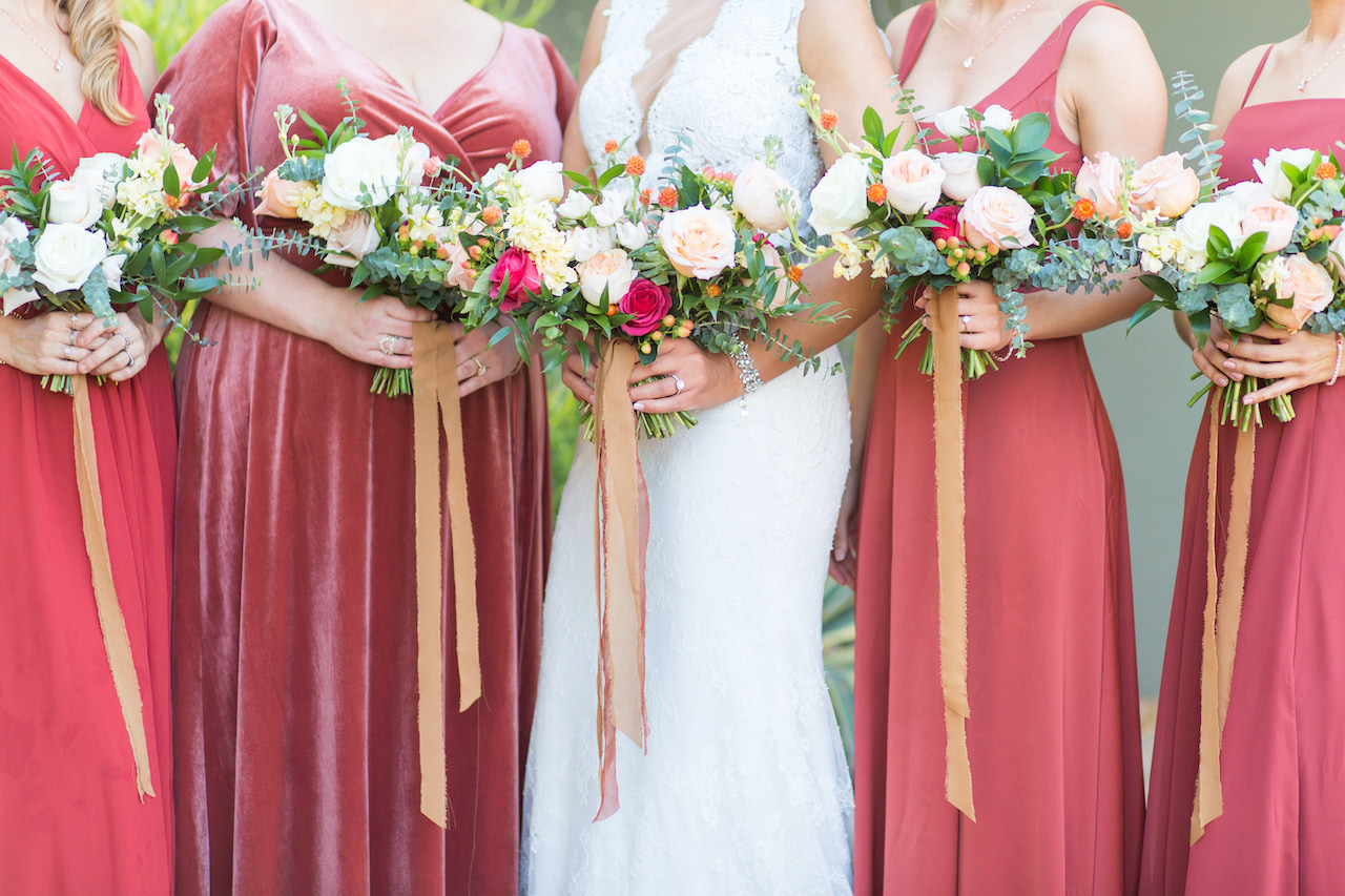 Bridesmaid dresses of rust red and bridal gown of white, holding bouquets.