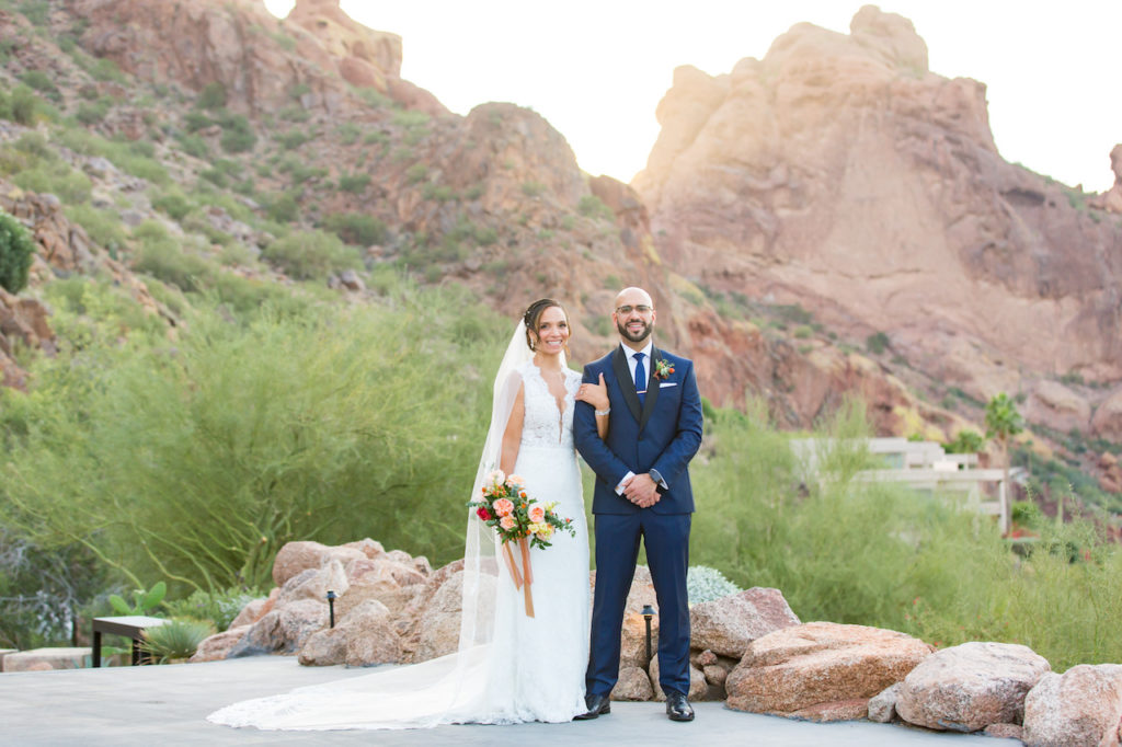 Bride and groom smiling, standing in front of desert landscape of mountains.