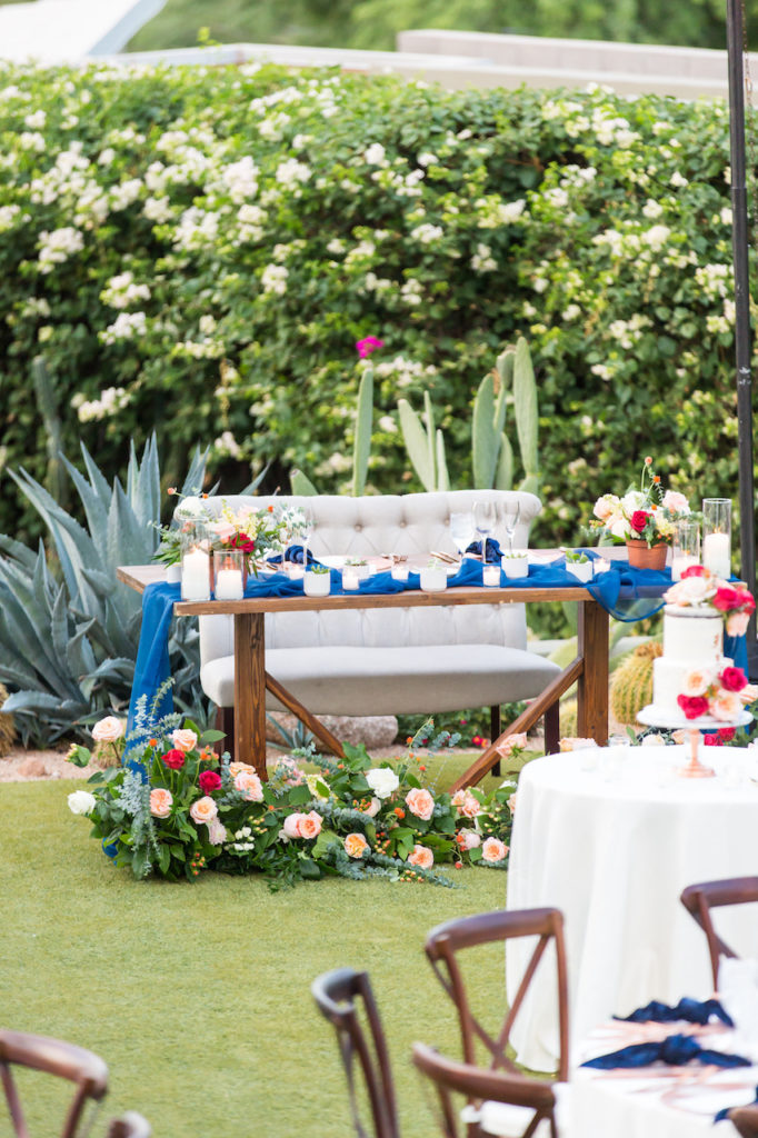 Sweetheart table with blue draping, floral on ground in front, floral arrangements and candles. Featuring a bench seat.