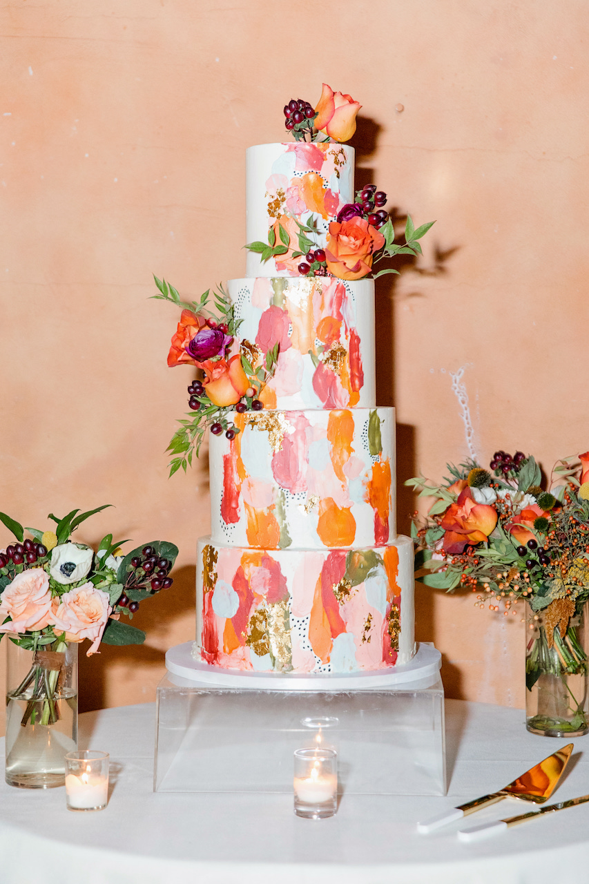 Bright four tiered wedding cake of white with painted pink, orange, gold and added matching flowers.