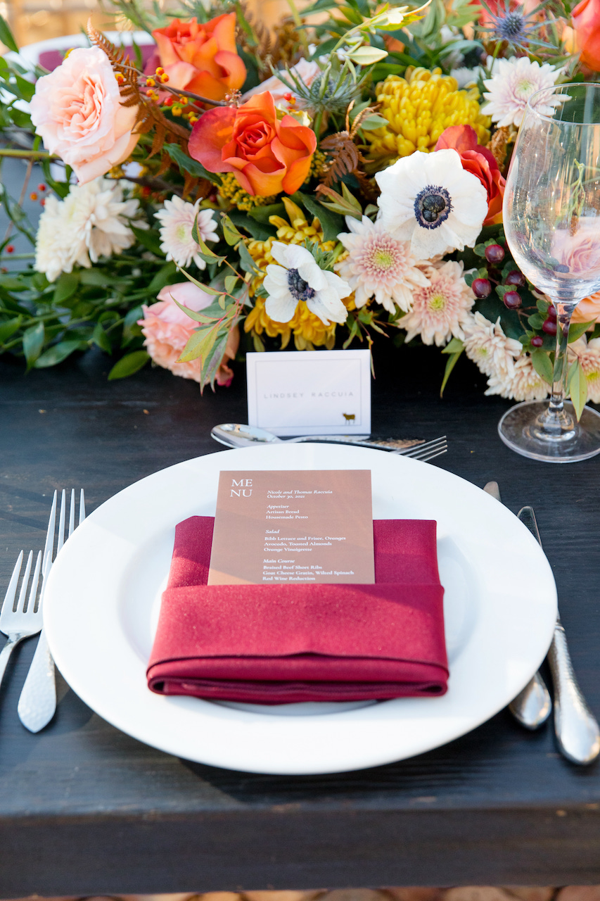 Fall wedding reception place setting with white plate, maroon napkin, and lush floral arrangement.