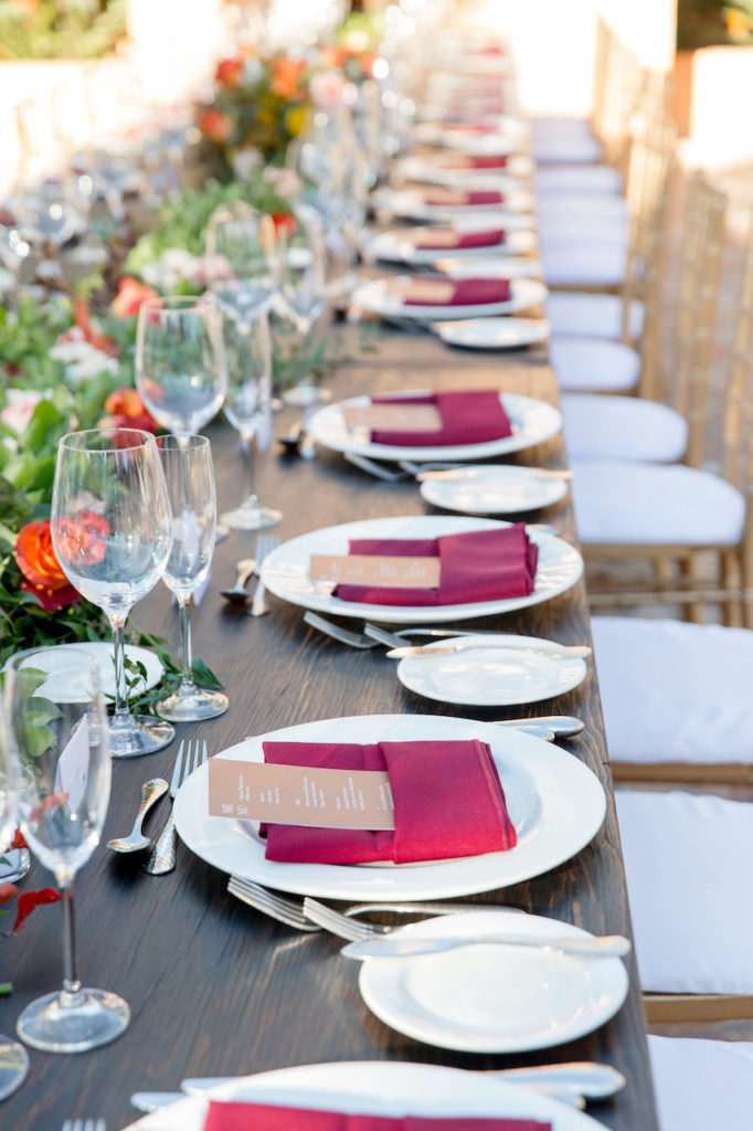 Long farm table at wedding reception with place setting of white plates, maroon napkins, menus.