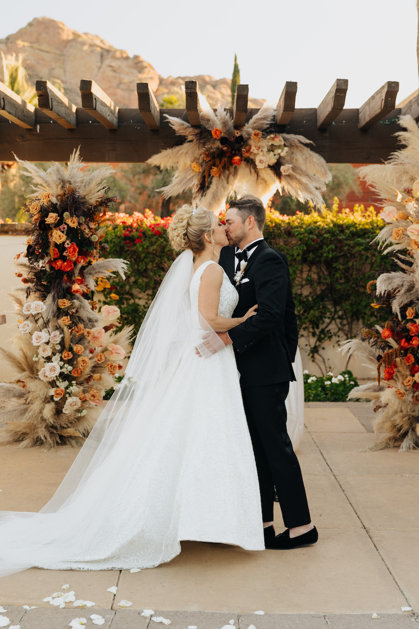Bride and groom kissing at wedding ceremony under pampas and flower installation.