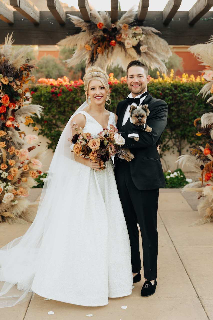 Bride and groom smiling, holding small dog in a tuxedo.