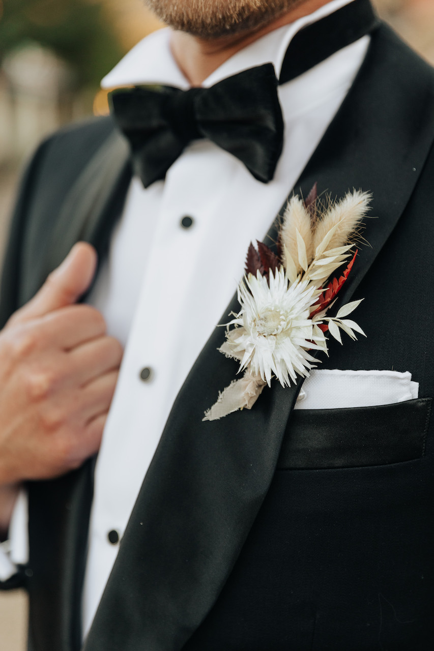 Groom boutonniere of dried floral elements in white and maroon/red.