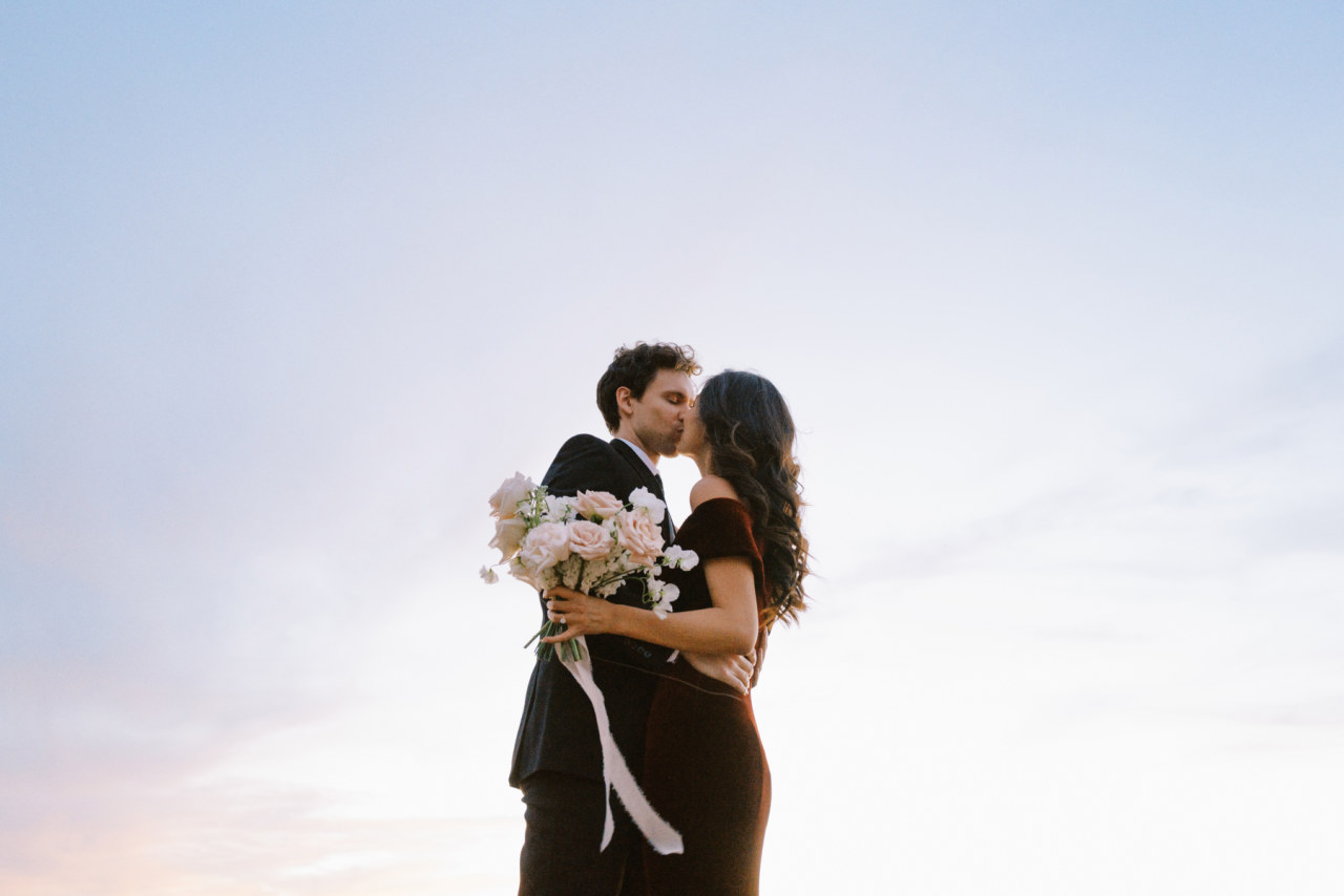 Woman holding bouquet and wearing red velvet dress kissing man in black suit.