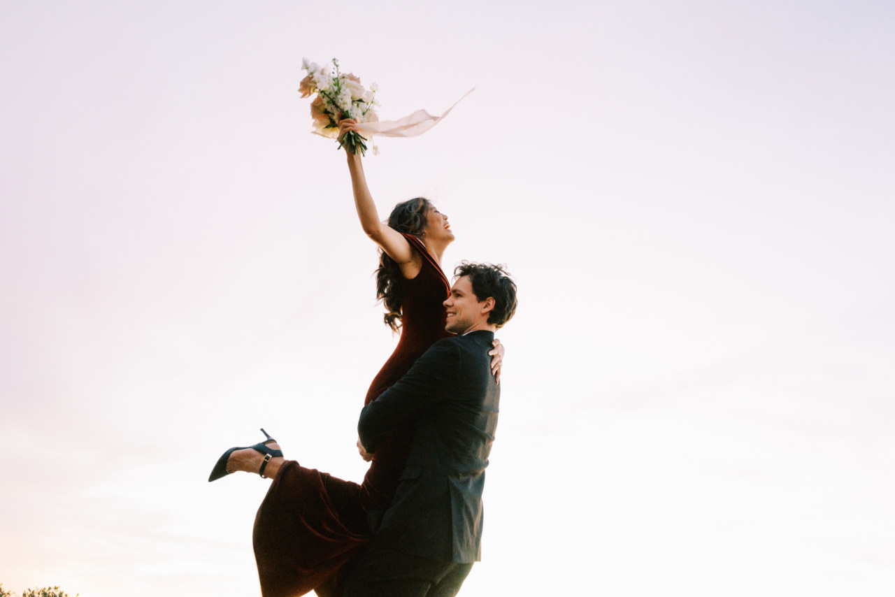 Man lifting up woman in hug while woman hold floral bouquet up in air.