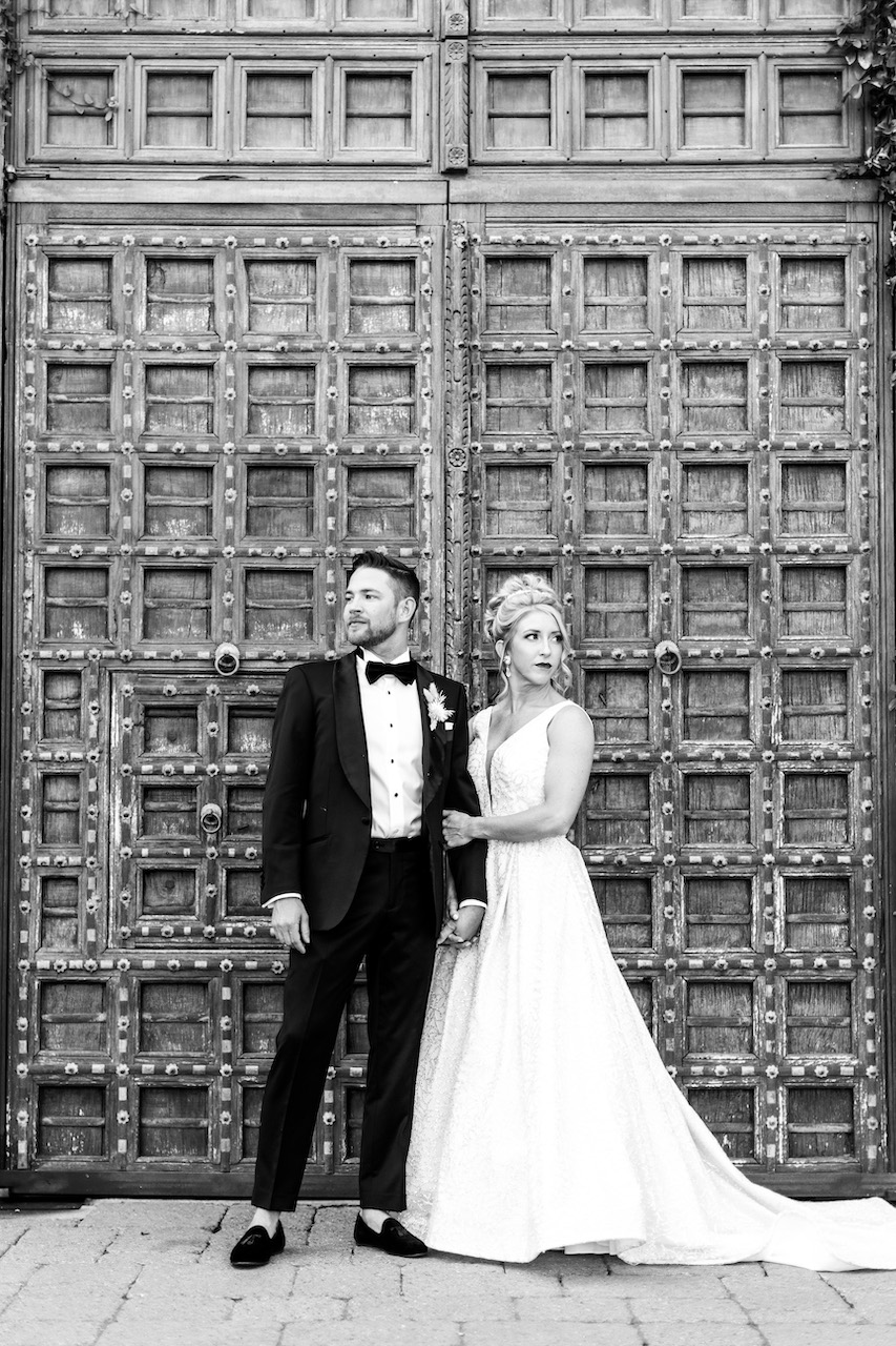 Bride and groom standing in front of large detailed wood doors, looking off into distance.