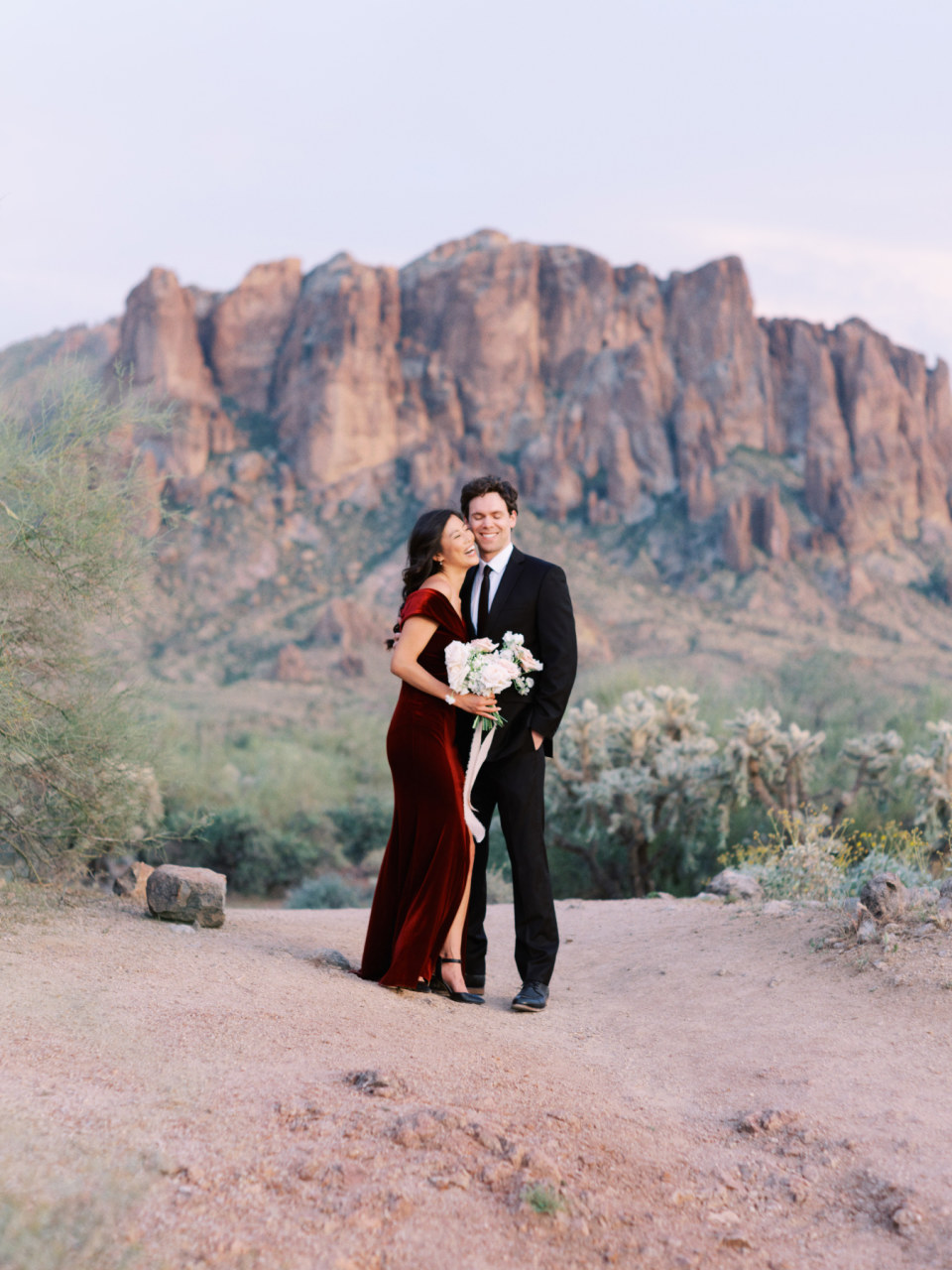 Woman and man smiling, embracing in desert landscape with Superstition Mountains in background.