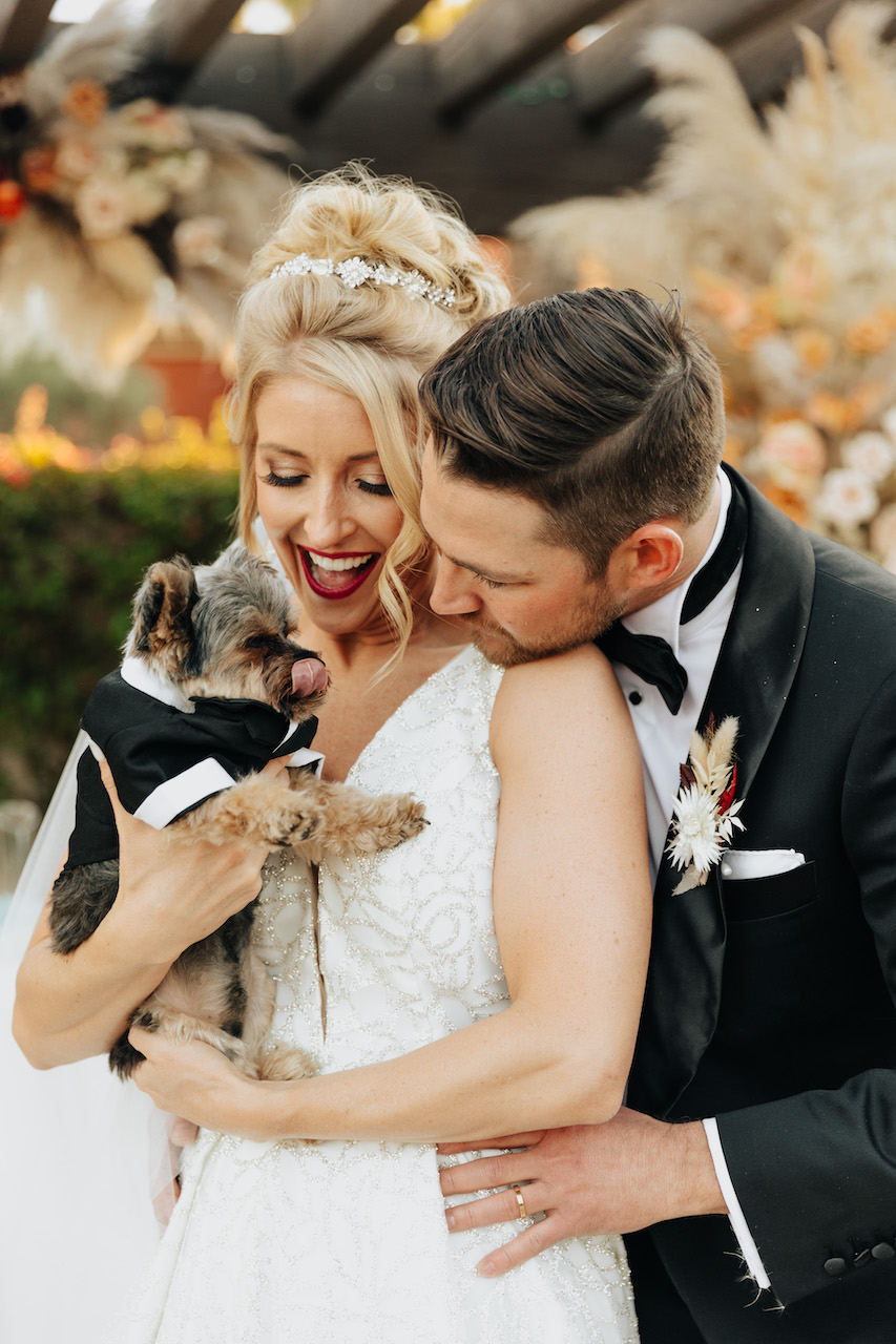 Bride and groom holding small dog in tuxedo, smiling.