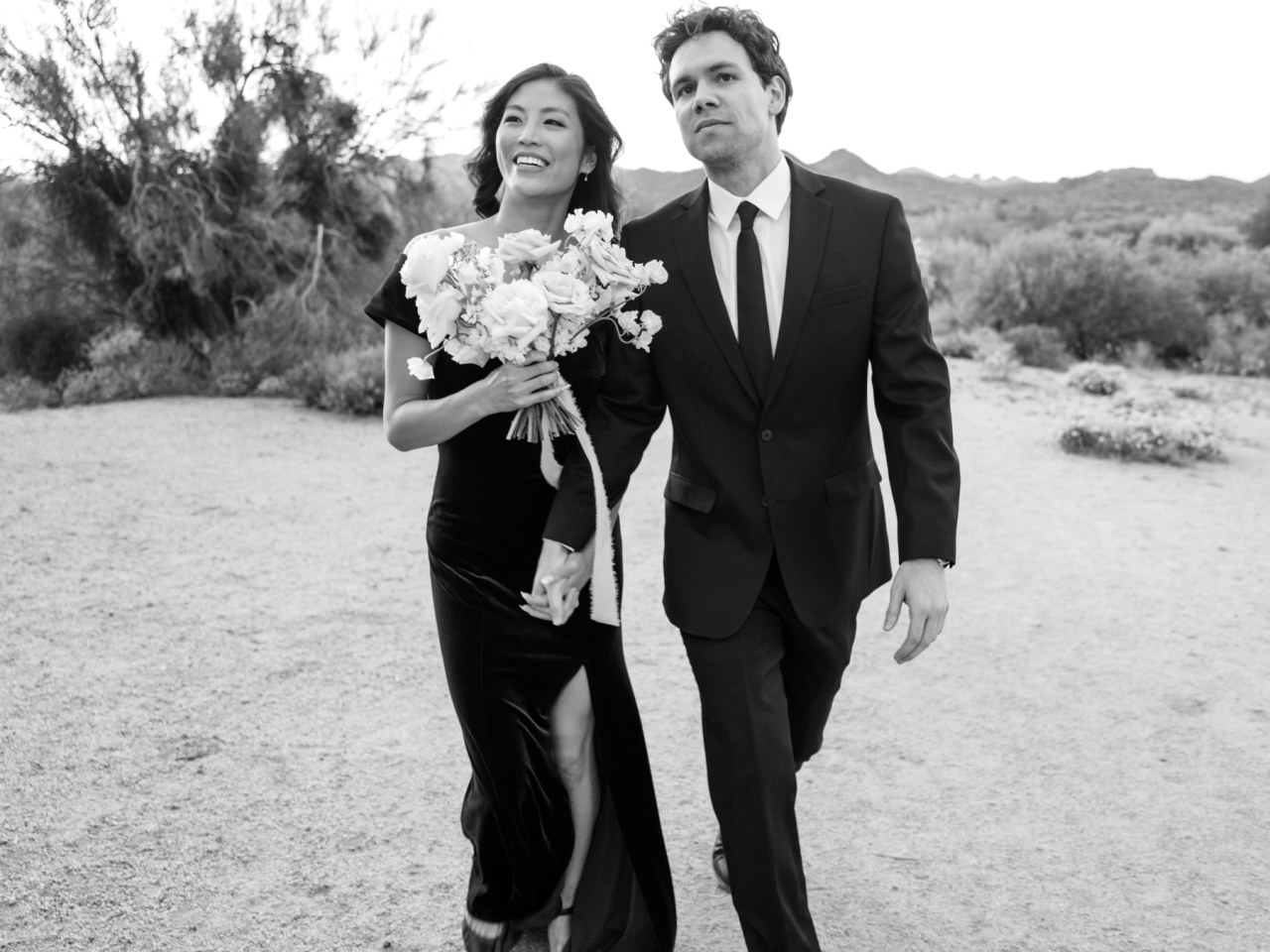 Man and woman holding hands walking in desert landscape. Woman holding bouquet.