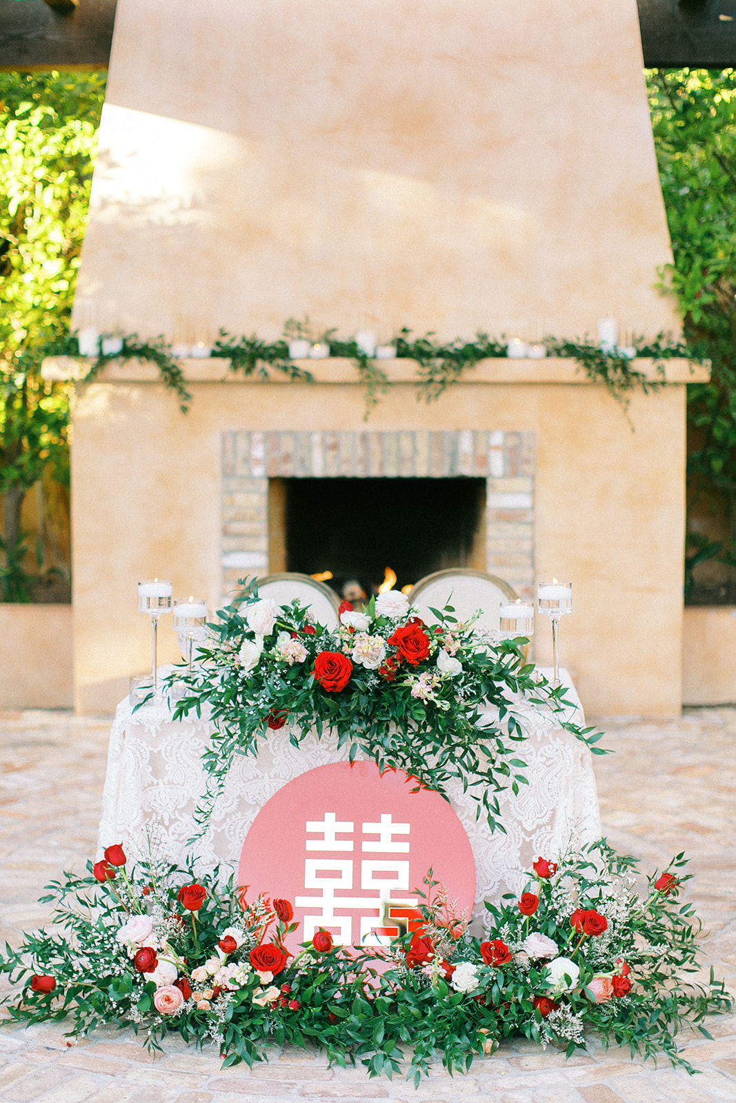 Sweetheart table floral arrangements of greenery and lush red, white, and pink roses on table and ground.