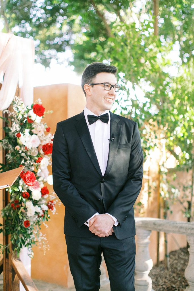 Groom in black tux standing at front of wedding ceremony.f