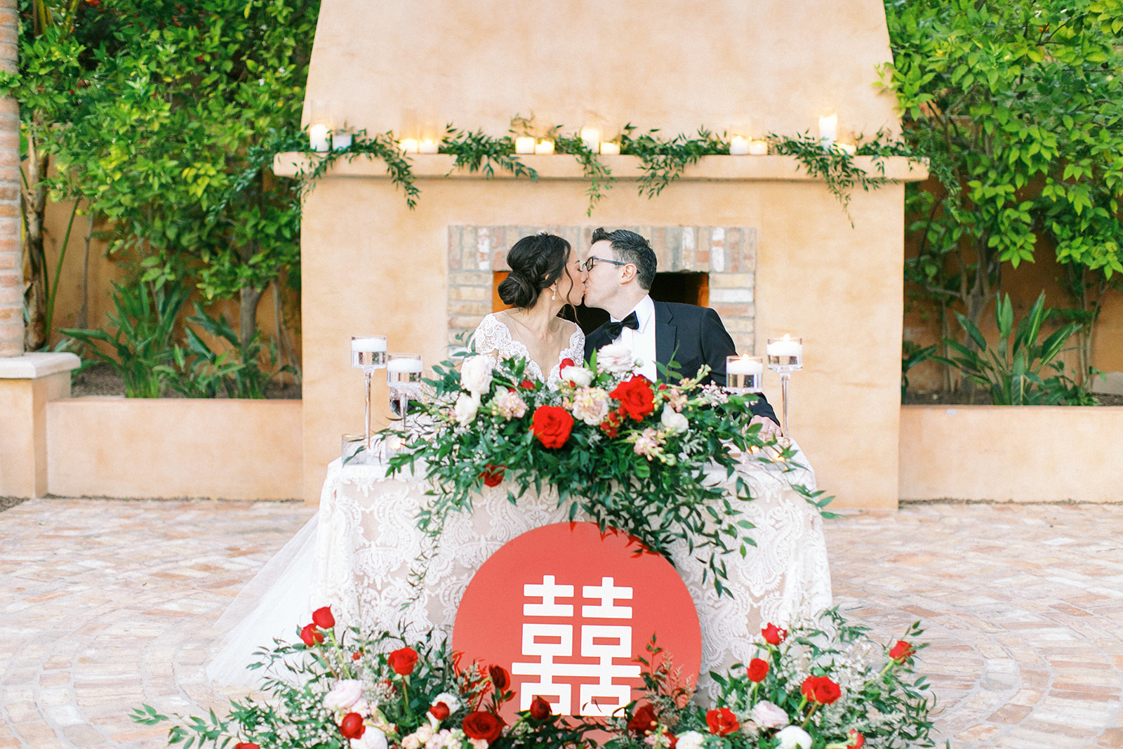 Bride and groom kissing at sweetheart table with floral and greenery arrangements and candles.