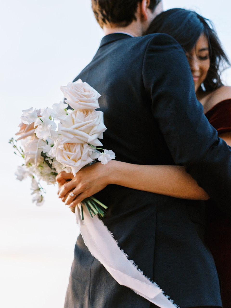 Woman and man hugging with woman's arms around his back, holding bouquet.
