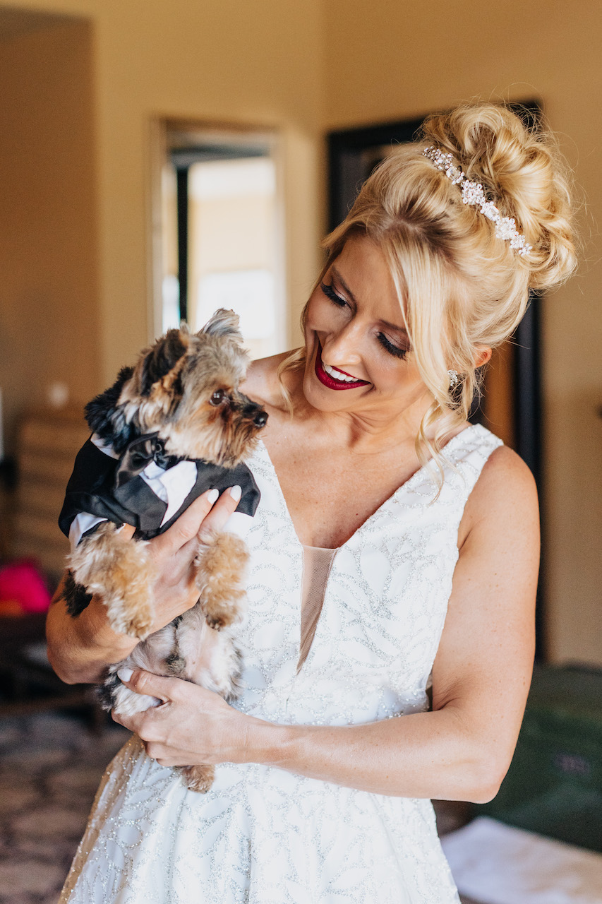 Bride smiling, holding small dog wearing a tuxedo.