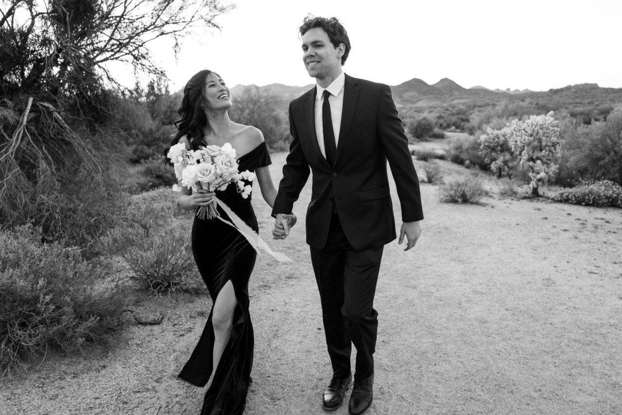 Man and woman walking in desert holding hands, woman holding bouquet.