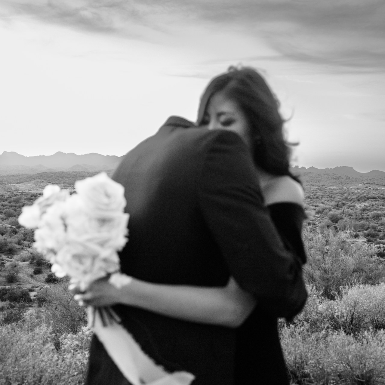 Woman and man hugging in desert landscape with woman holding bouquet.