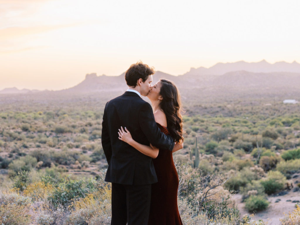 Woman with arm around man, kissing each other with Superstition Mountains in distance.