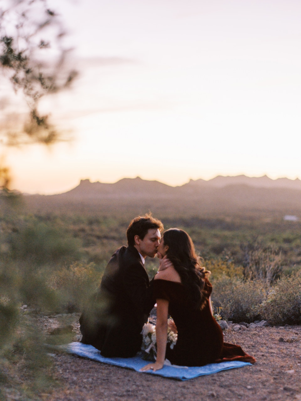 Couple sitting on blue blanket, kissing in Arizona desert landscape.