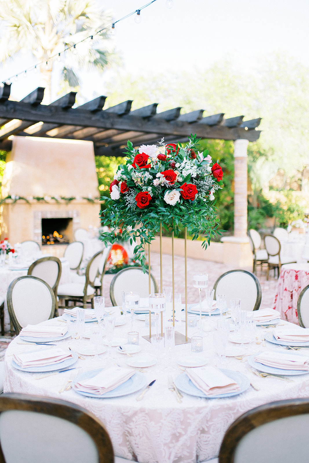 Tall brass wedding reception centerpiece of greenery and white, pink and red roses on round table.