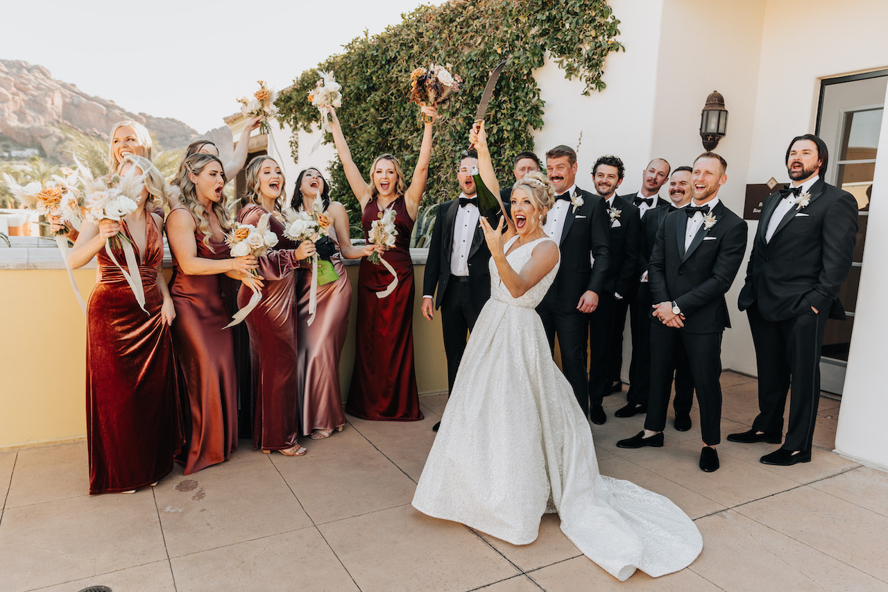 Bride opening a bottle of champagne with bridal party celebrating behind her.