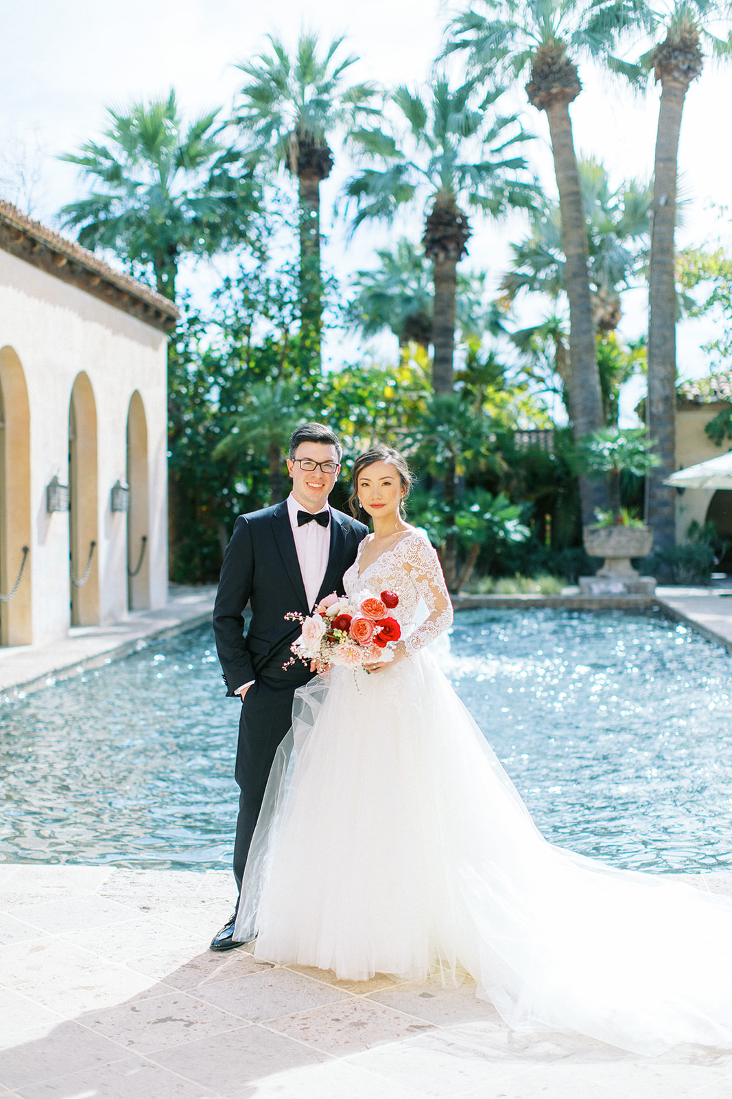 Groom and bride holding bouquet standing next to each other in front of large fountain pool.