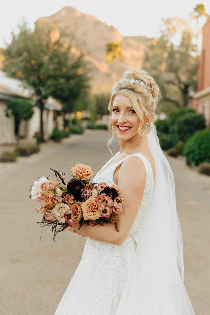 Bride smiling at camera, holding bouquet, standing outside in desert setting.