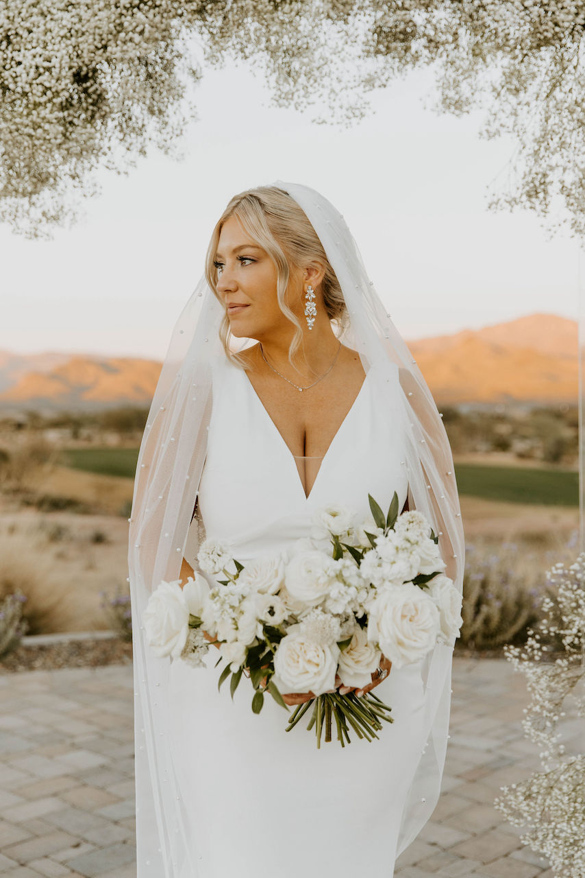 Bride holding white floral bouquet with long veil looking off to side under baby's breath arch.