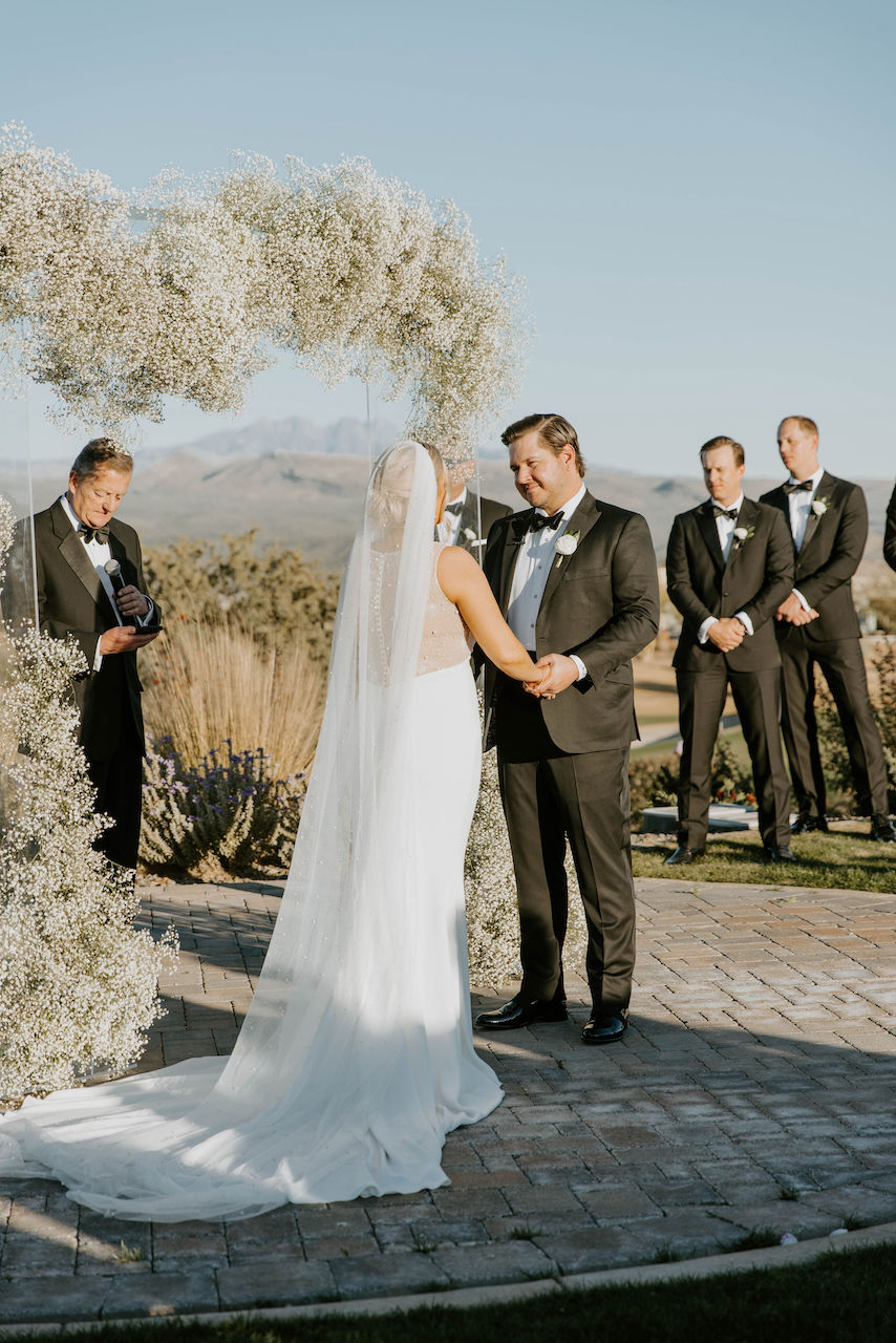 Bride and groom holding hands during outdoor wedding ceremony in desert landscape with baby's breath arch.