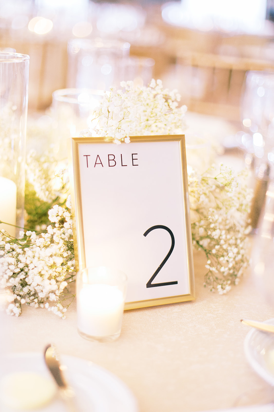 Gold framed table number sign with black font and candles with baby's breath behind it.