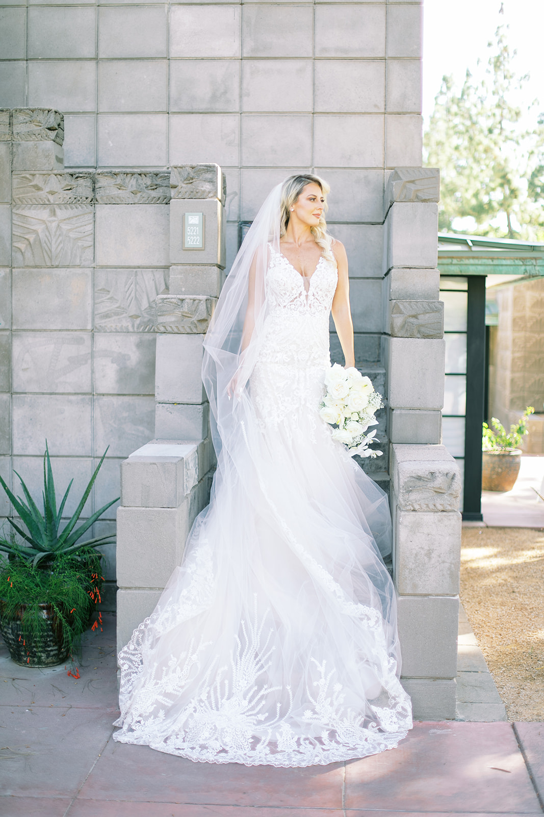 Bride standing on stairs of gray building holding out veil and bouquet, looking to side.