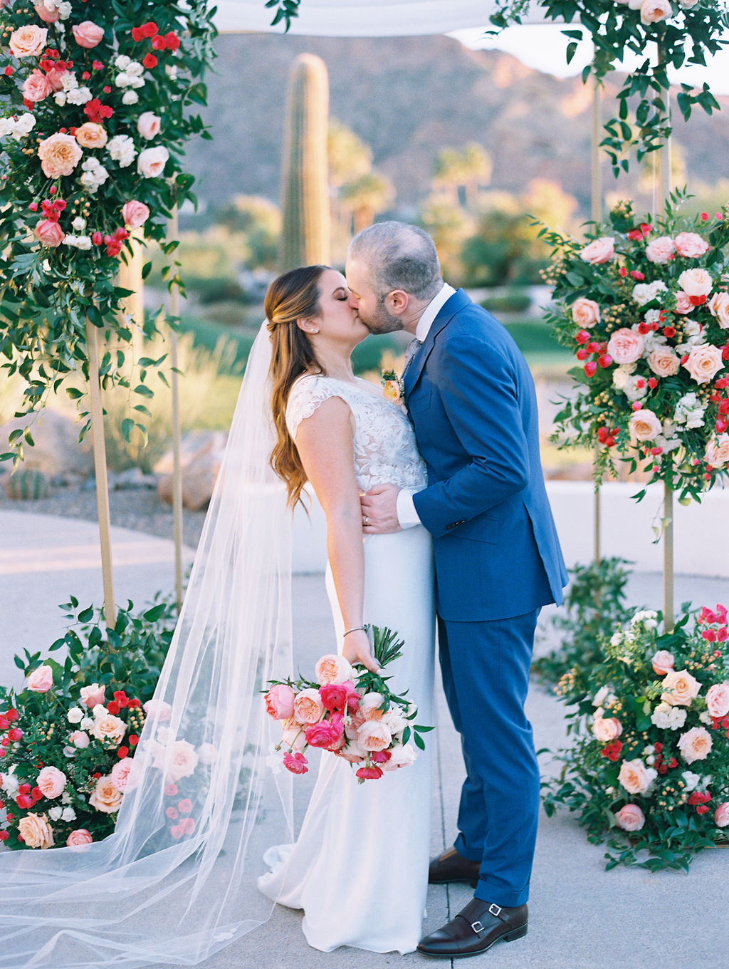Bride and groom kissing under chuppah with floral installations of pink and peach flowers.