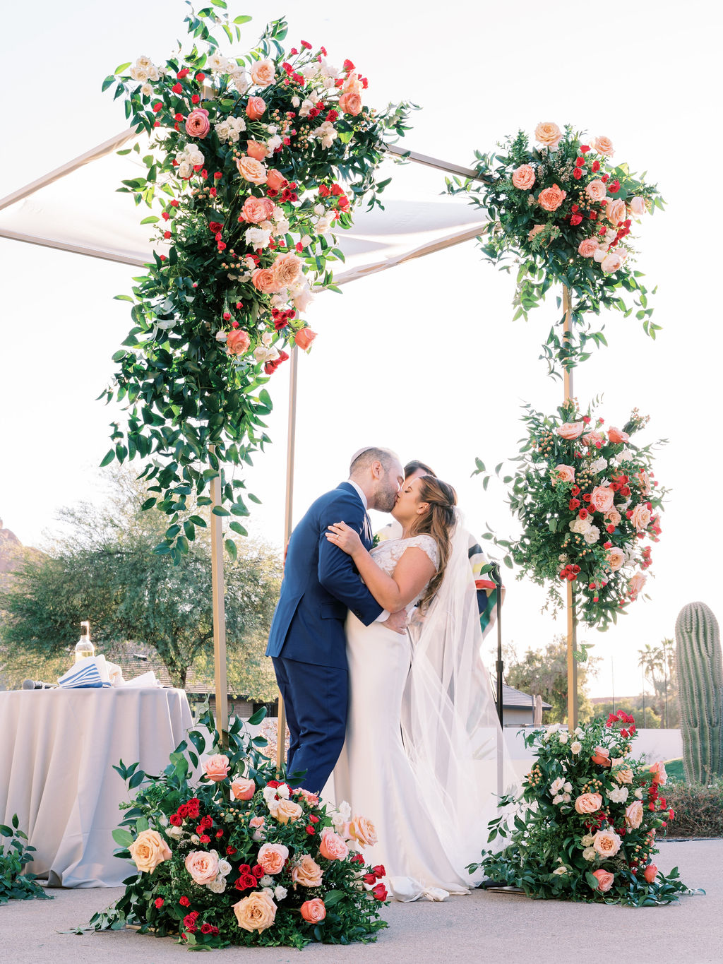 Bride and groom kissing under chuppah with floral installations of pink and peach flowers.