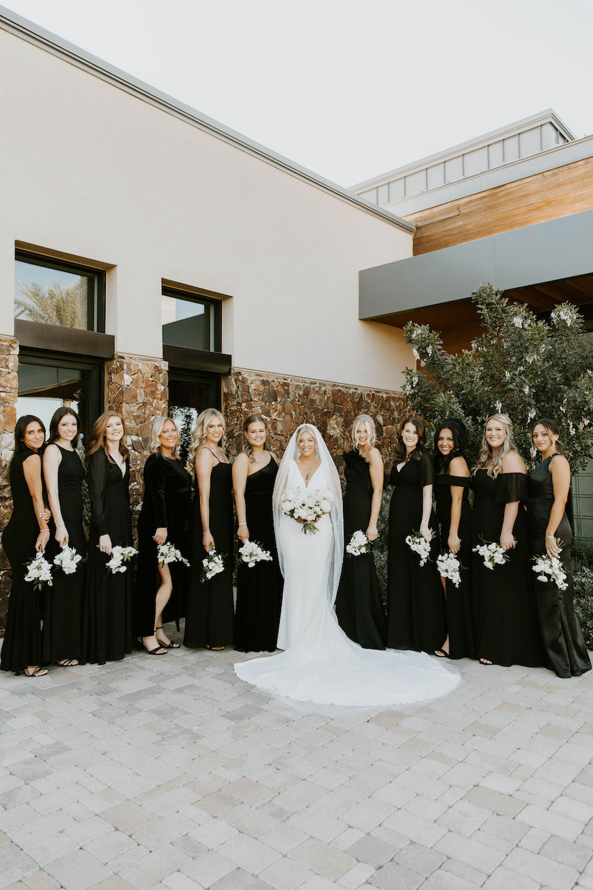 Bride standing in middle of line of bridesmaids wearing black dresses and holding small white flowers bouquets.
