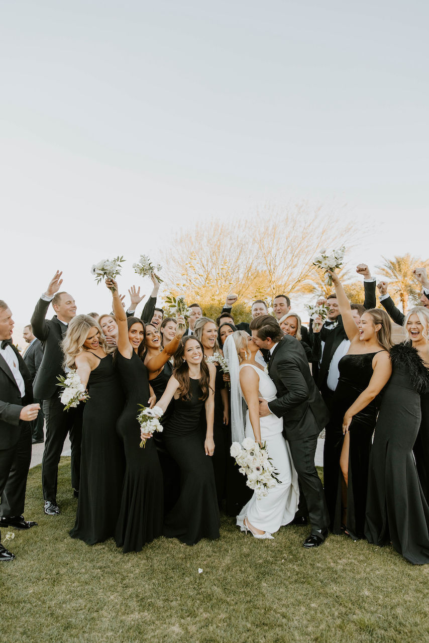 Wedding party wearing black celebrating while bride and groom kiss in center.