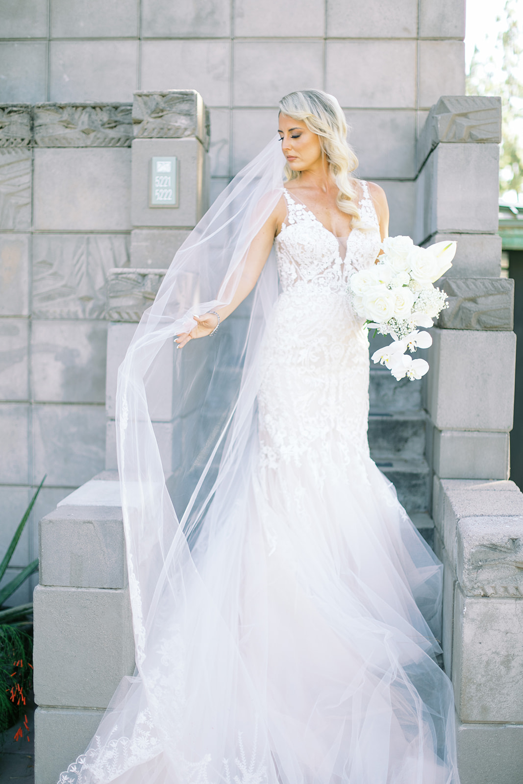 Bride standing on stairs of gray building holding out veil and bouquet, looking to side.