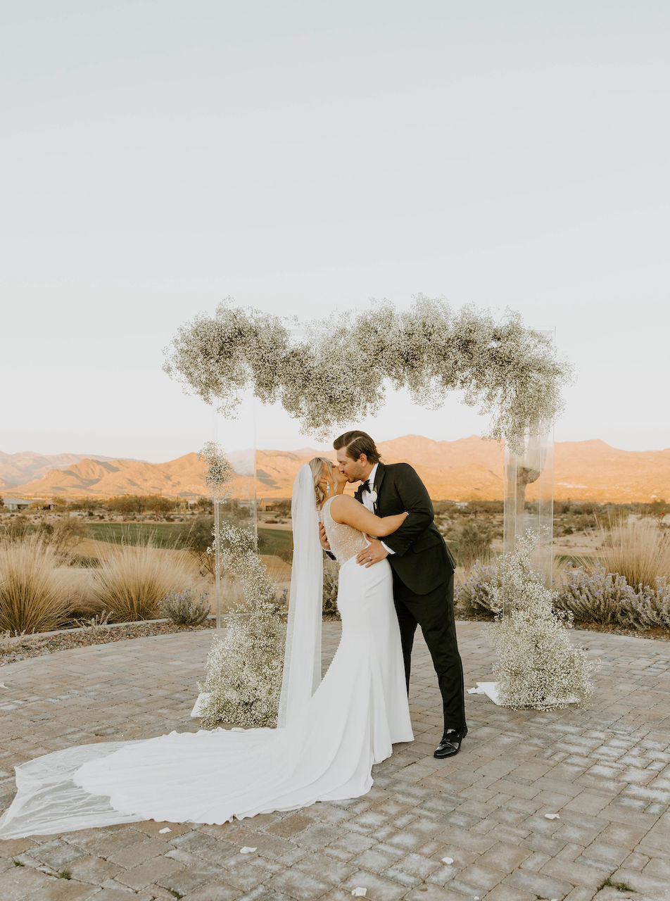 Bride and groom kissing outside in front of desert landscape with baby's breath on clear acrylic arch.