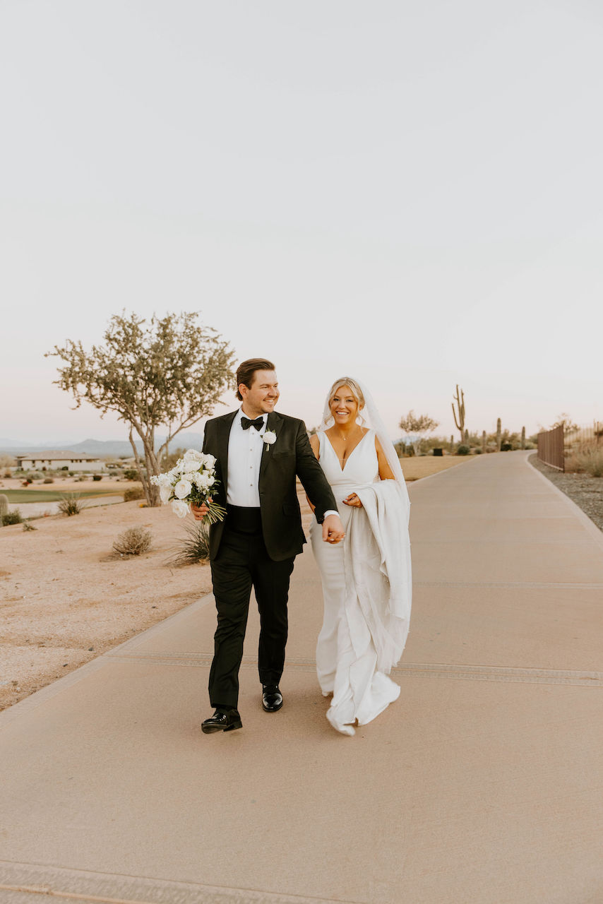 Bride and groom walking down sidewalk holding hands in desert setting.