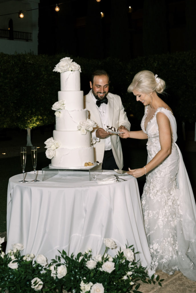 Bride and groom cutting five tiered white wedding cake with white roses.