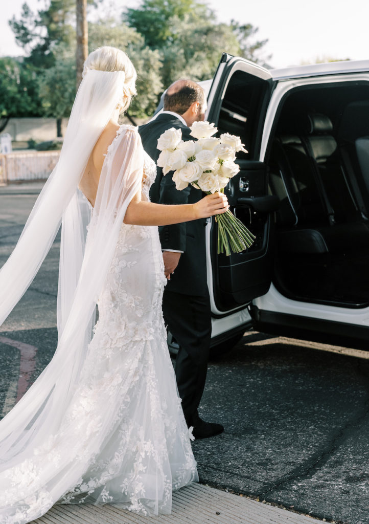 Bride and groom exiting into SUV with bride holding long stem white roses bouquet.
