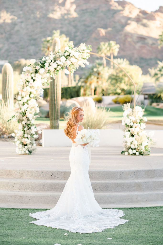Bride looking over shoulder at bouquet she is holding at the base of stairs of outdoor wedding ceremony stage with arch.