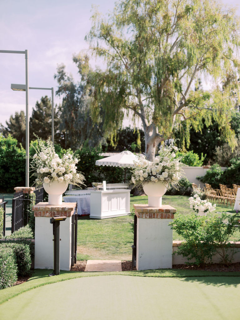 Ceremony entrance with two large white floral and greenery arrangements in large vases on low brick pillars on both sides of entrance.