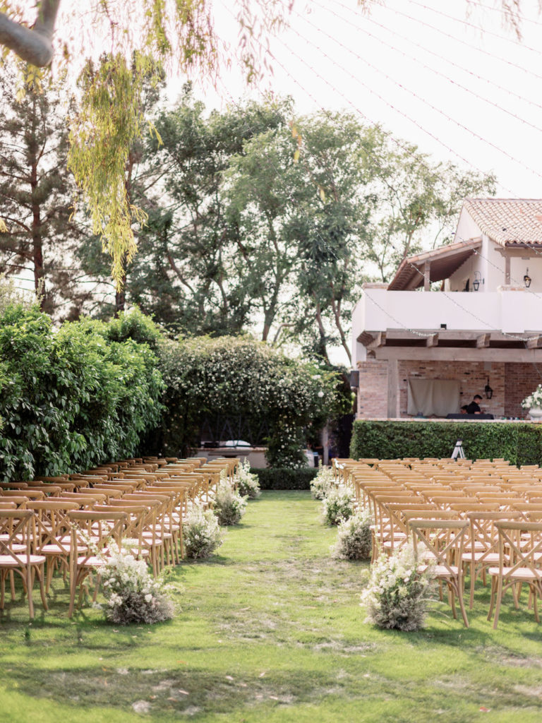 Outdoor wedding ceremony on back lawn of home with wood chairs and aisle floral arrangements.