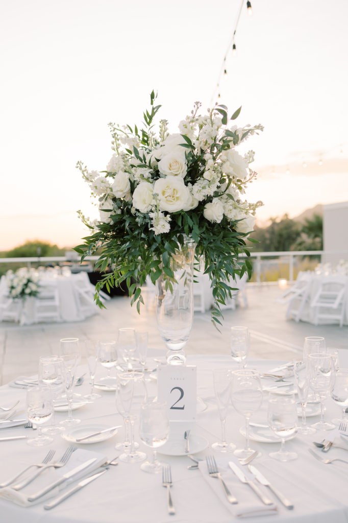 Tall wedding reception centerpiece in clear glass vase of white flowers and greenery.