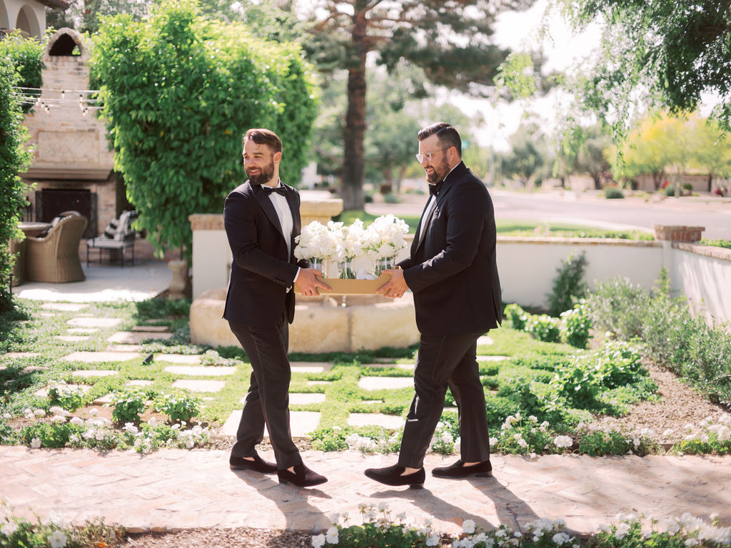 Groomsmen in black suits walking while holding a box of bouquets in glass vases.