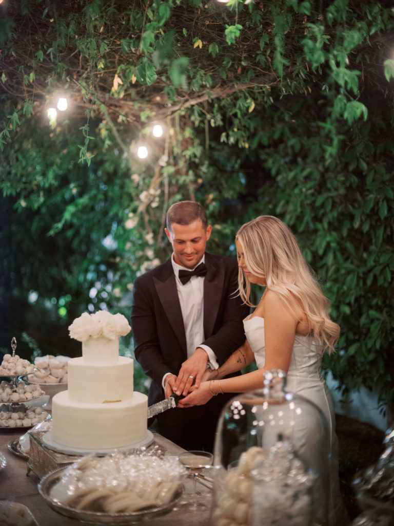 Bride and groom cutting three tiered white wedding cake with greenery canopy around them.