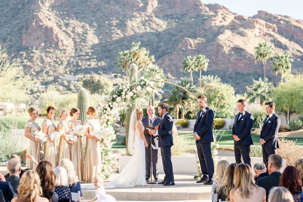 Front of wedding ceremony with groom and bride standing with officiant and bridesmaids and groomsmen standing to side. Gold wedding arch and mountain in distance.