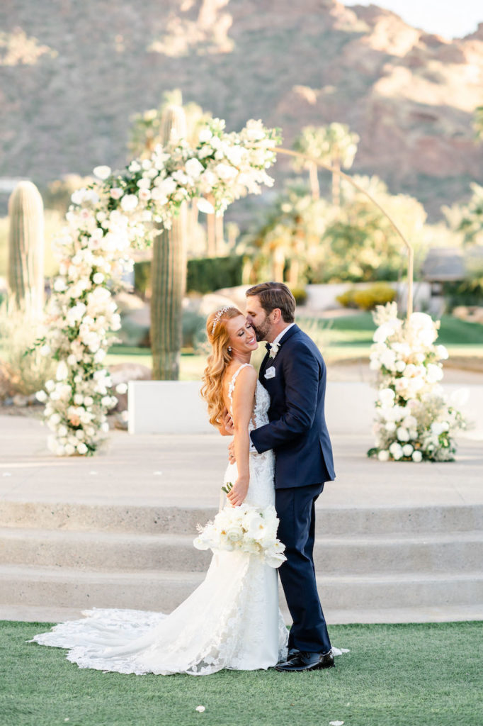 Groom hugging bride, kissing her cheek in outdoor in front of gold arch with white floral and greenery installed on it.
